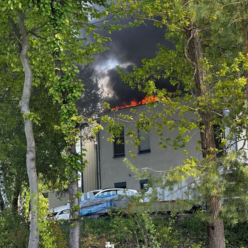 Ein Gebäude mit einem Feuer auf dem Dach ist von grünen Bäumen und geparkten Autos umgeben. Der Rauch ist dunkel und dick.