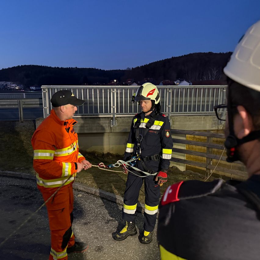 Drei Feuerwehrleute in Uniform bereiten sich auf einen Rettungseinsatz im Freien vor. Einer hält ein Seil. Hinter ihnen ist ein Metallzaun und eine hügelige Landschaft zu sehen.