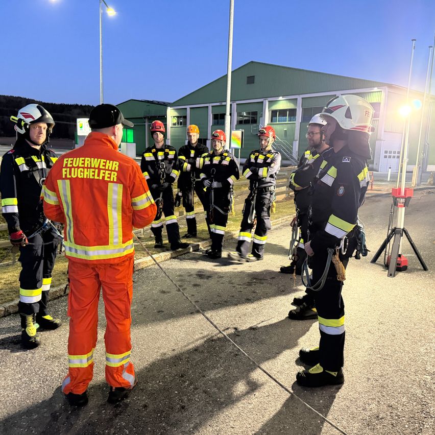 Eine Gruppe Feuerwehrleute in voller Ausrüstung stehen draußen versammelt, wahrscheinlich in Vorbereitung auf eine Trainingseinheit. Sie tragen Helme und orange Uniformen.