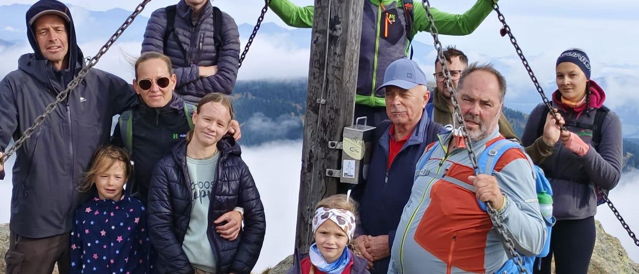 Eine Gruppe von Wanderern steht auf einem Berggipfel, einige halten sich an Ketten fest und andere halten Stöcke. Sie tragen alle Jacken, einige mit Kapuzen. Die Gruppe besteht aus Kindern und Erwachsenen, alle lächeln.