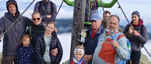 Eine Gruppe von Wanderern steht auf einem Berggipfel, einige halten sich an Ketten fest und andere halten Stöcke. Sie tragen alle Jacken, einige mit Kapuzen. Die Gruppe besteht aus Kindern und Erwachsenen, alle lächeln.