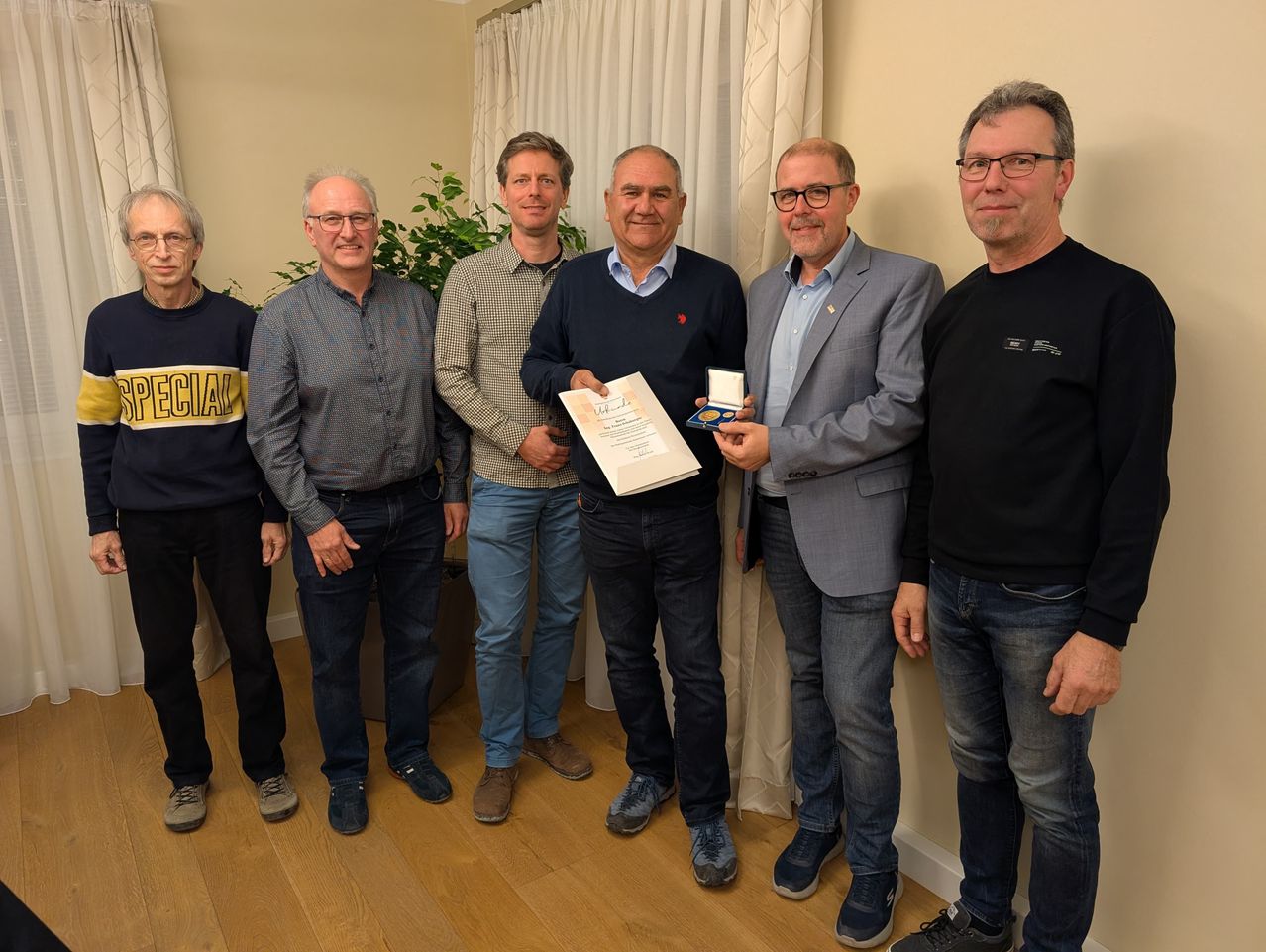 Eine Gruppe von Männern steht in einem Raum, einer hält ein Papier und eine Medaille. Sie tragen Freizeitkleidung und Brillen. Eine Pflanze und Vorhänge sind im Hintergrund.