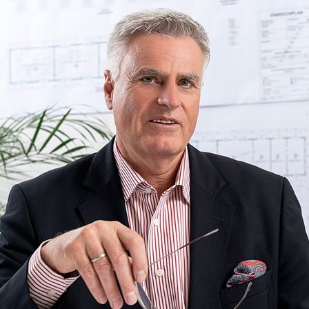 Ein Mann im Anzug hält eine Brille in der Hand, er sitzt vor Architekturplänen.