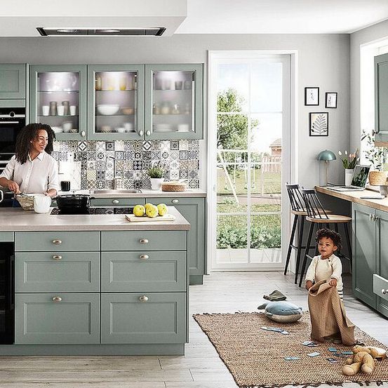 Eine Frau bereitet in einer Küche mit grauen Schränken und Arbeitsplatten Essen zu. Ein Kind steht in der Nähe auf einem Teppich. Die Küche hat ein Fenster und eine Tür, die auf eine Terrasse führt.