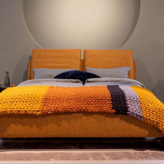 Ein modernes Schlafzimmer mit einem Doppelbett mit orangefarbenen Kopfteilen und einer farbenfrohen, strukturierten Decke. Kissen sind ordentlich platziert, und eine kleine Flasche steht auf dem Nachttisch. Der Raum hat ein sauberes, minimalistisches Design.