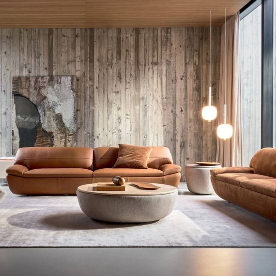 Ein modernes Wohnzimmer mit Holzwänden, zwei braunen Ledersofas, einem runden Couchtisch und hängenden Lampen.