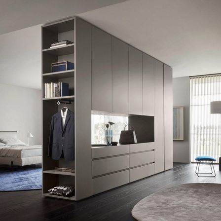 Ein modernes Schlafzimmer mit einem großen grauen Kleiderschrank, einem Bett und einem Teppich. Der Kleiderschrank hat Regale, Schubladen und einen Aufhängebereich. Eine Handtasche und Bücher sind auf dem Regal.