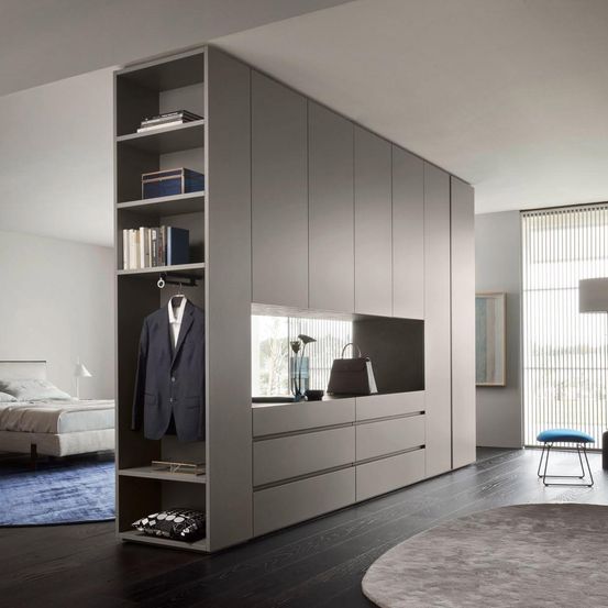 Ein modernes Schlafzimmer mit einem großen grauen Kleiderschrank, einem Bett und einem Teppich. Der Kleiderschrank hat Regale, Schubladen und einen Aufhängebereich. Eine Handtasche und Bücher sind auf dem Regal.