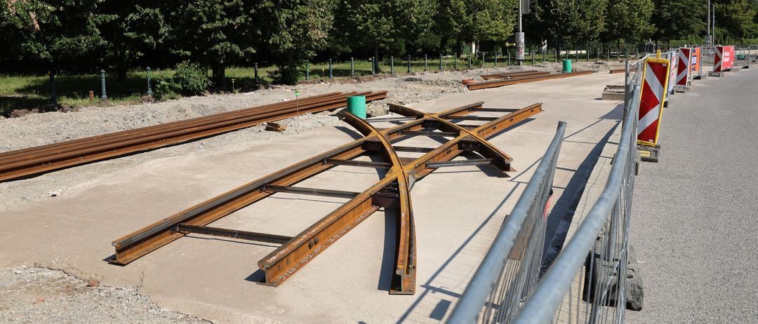 Ein Bereich mit Bauarbeiten an Eisenbahnschienen, verlegten Metallschienen, Bäumen im Hintergrund und einer Straßenlaterne rechts.