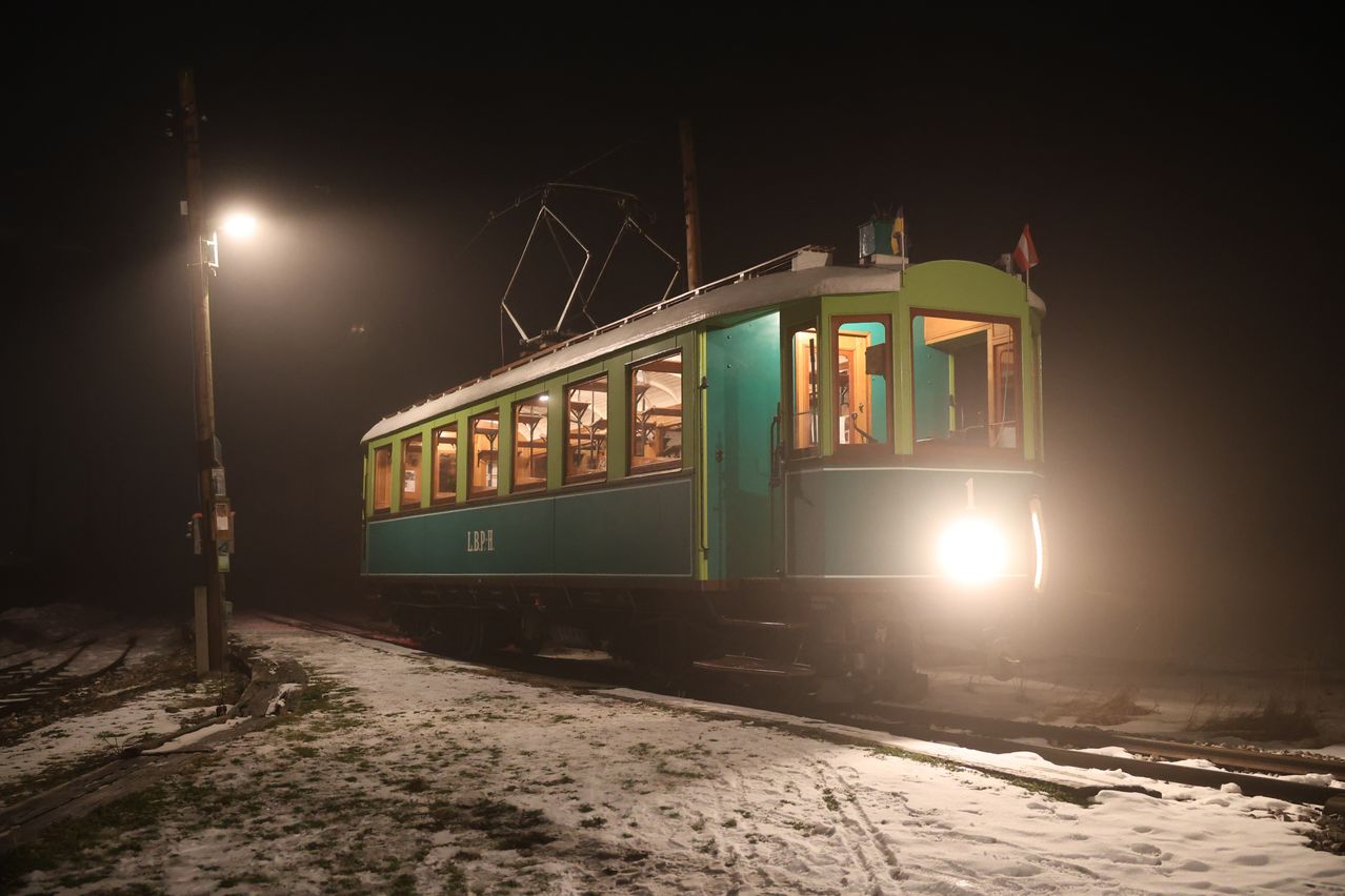 Ein grüner Vintage-Zug mit beleuchteten Fenstern fährt bei Nacht auf den Schienen, beleuchtet von einer Straßenlaterne. Der Boden ist schneebedeckt und eine Flagge ist am Zug sichtbar.