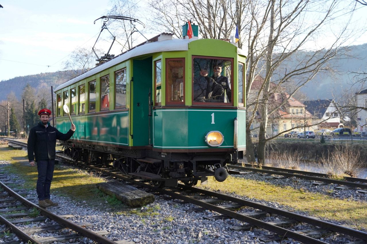 Ein grüner altertümlicher Zug mit der Nummer 1 an der Front, gefahren von zwei Männern, steht auf den Gleisen in der Nähe eines Flusses mit kahlen Bäumen.