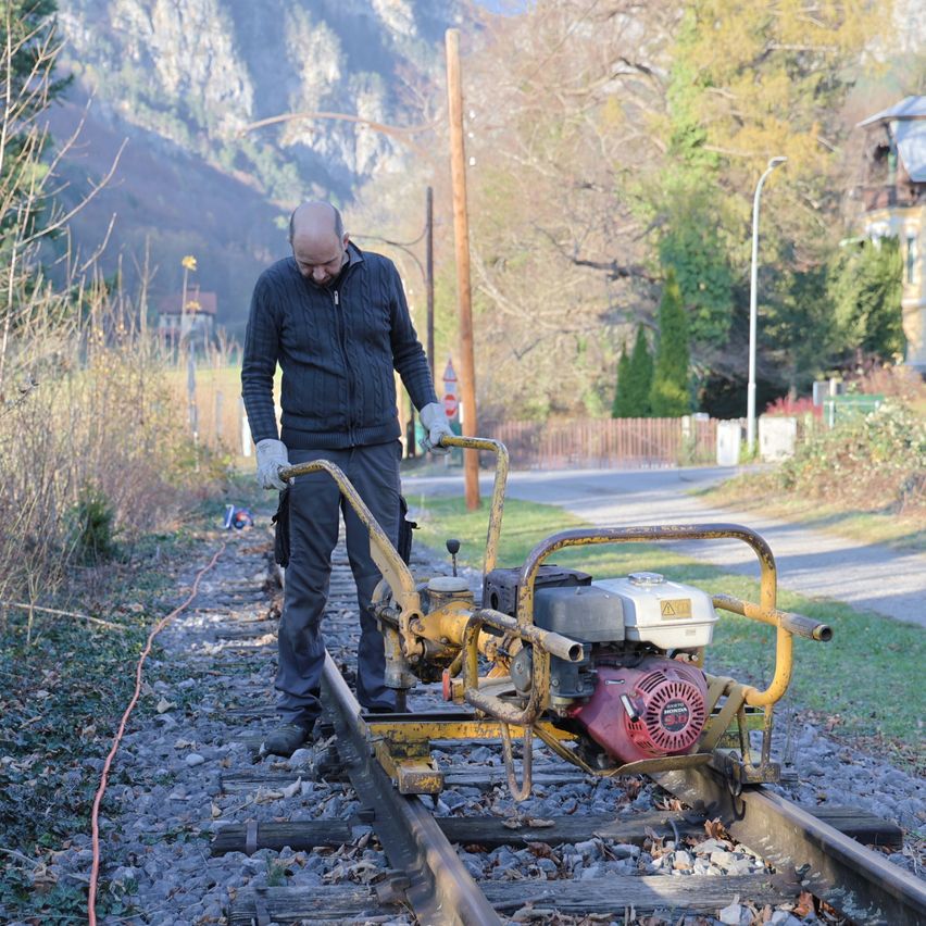 A man is working on a railway track, using a yellow machine with a red engine. He is standing on the tracks, and there are trees and a house in the background.