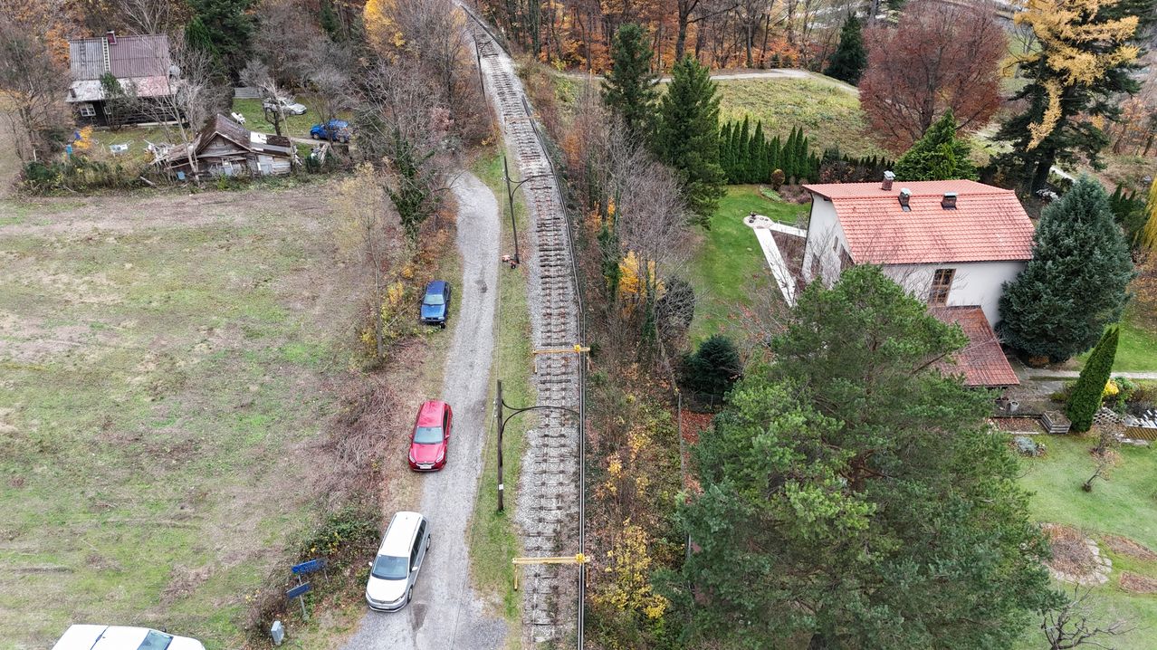 An aerial view of a narrow gravel road with parked cars, one blue and one red, along the side. The road runs parallel to a railroad track with multiple train tracks and trees on both sides. A house with a red roof is visible on the right.