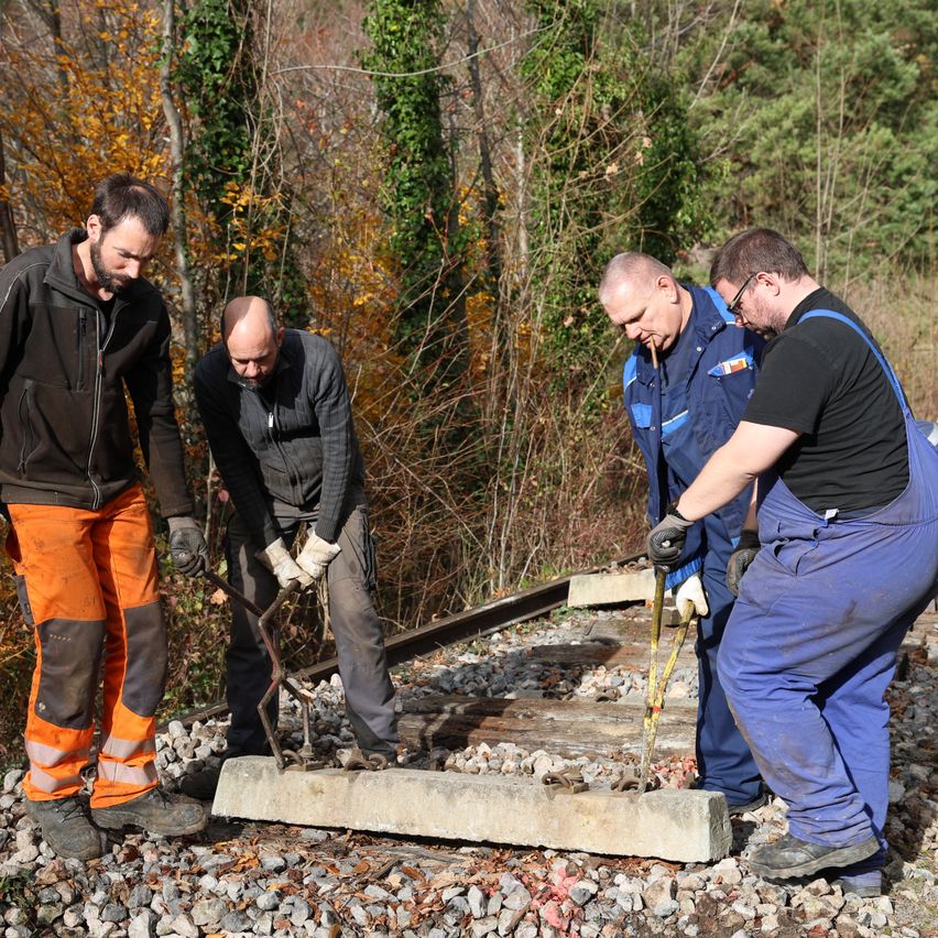 Four men in work attire are working on a railway track, surrounded by trees and gravel. One holds a tool, another smokes a cigar.