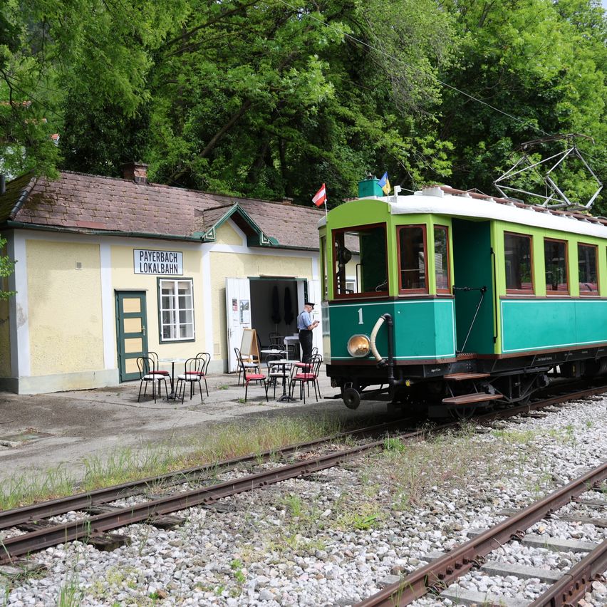 Ein grüner Straßenbahnwagen ist am Bahnhof Payerbach Lokalbahn geparkt, wobei ein Mann außerhalb des Bahnhofsgebäudes steht und Tische und Stühle auf dem Bahnsteig aufgestellt sind.