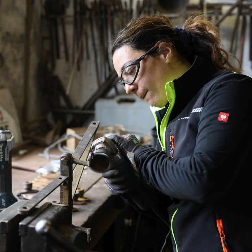 Eine Frau mit Schutzbrille und Warnweste arbeitet in einer Werkstatt und bearbeitet ein Metallobjekt mit Werkzeugen.