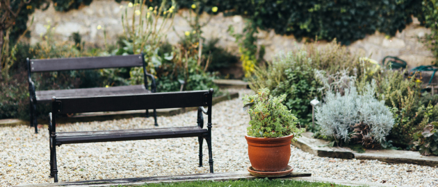 Ein kleiner Garten mit Kieswegen, zwei Bänken und einer Topfpflanze, umgeben von grünem Laub und einer Steinmauer.