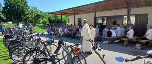 Ein Priester steht vor einer Gruppe von Menschen, die auf Bänken und Stühlen vor einem Gebäude sitzen, mit geparkten Fahrrädern in der Nähe.