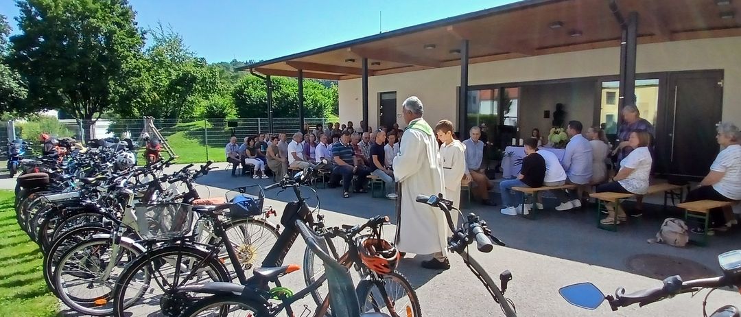 Ein Priester steht vor einer Gruppe von Menschen, die auf Bänken und Stühlen vor einem Gebäude sitzen, mit geparkten Fahrrädern in der Nähe.