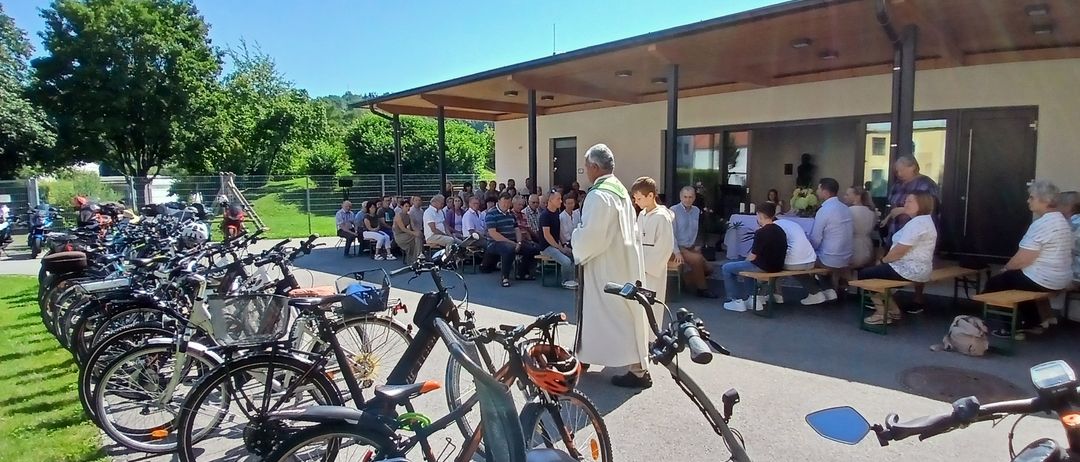 Ein Priester steht vor einer Gruppe von Menschen, die auf Bänken und Stühlen vor einem Gebäude sitzen, vor dem Fahrräder geparkt sind.