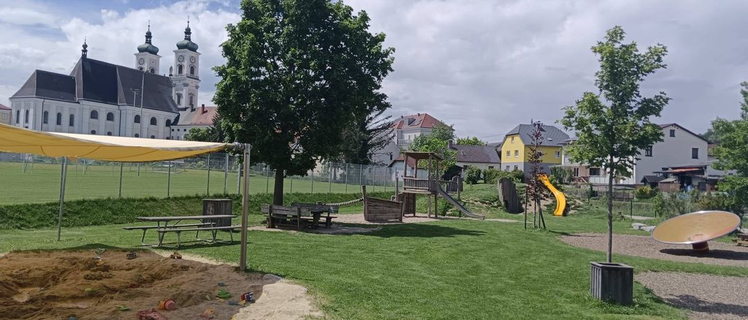 Ein Spielplatz mit einem gelben Rutschen und einem Picknickbereich befindet sich unter einem Baum. Es gibt ein gelbes Vordach, einen Zaun und ein Gebäude im Hintergrund.