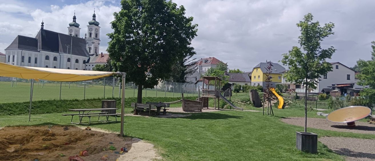 Ein Spielplatz mit einem gelben Rutschen und einem Picknickbereich befindet sich unter einem Baum. Es gibt ein gelbes Vordach, einen Zaun und ein Gebäude im Hintergrund.