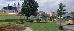 Ein Spielplatz mit einem gelben Rutschen und einem Picknickbereich befindet sich unter einem Baum. Es gibt ein gelbes Vordach, einen Zaun und ein Gebäude im Hintergrund.