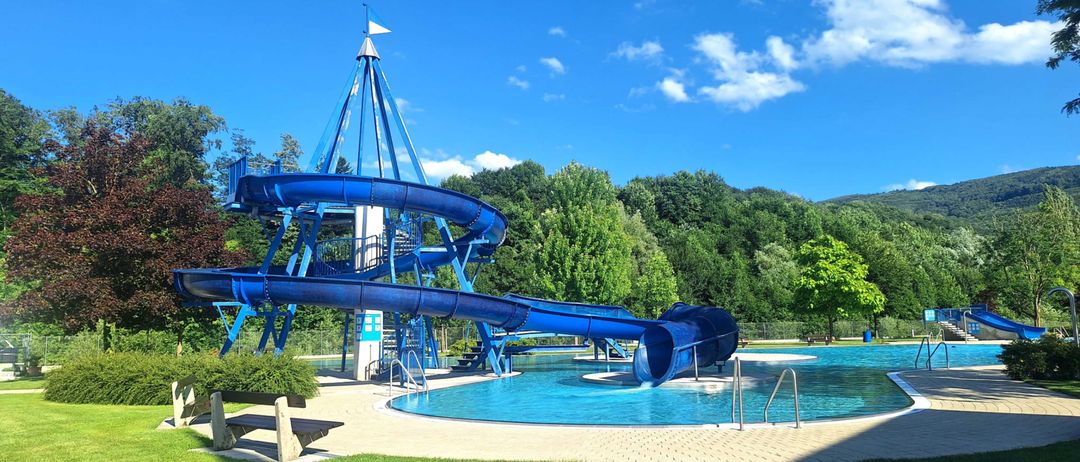 Eine große blaue Wasserrutsche mit einem Turm oben befindet sich in einem Poolbereich, umgeben von Bäumen und einem klaren blauen Himmel.