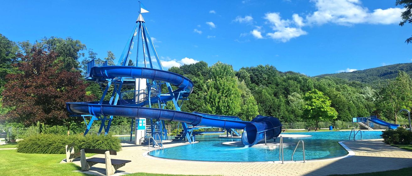 Eine große blaue Wasserrutsche mit einem Turm oben befindet sich in einem Poolbereich, umgeben von Bäumen und einem klaren blauen Himmel.