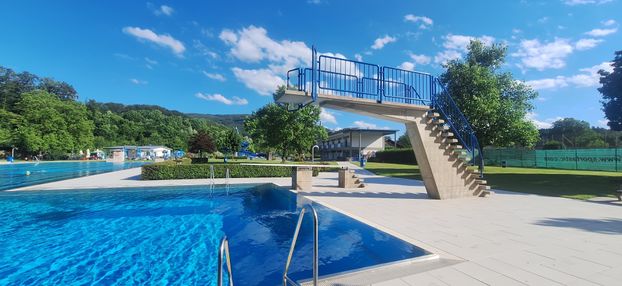 Ein blauer Sprungturm über einem Schwimmbecken mit klarem Wasser und blauen Geländern. Bäume und ein Gebäude sind im Hintergrund zu sehen.