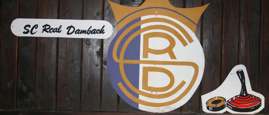 Eine hölzerne Wand zeigt ein Schild mit der Aufschrift 'Real Dambach' und ein Logo mit Krone und den Buchstaben 'RD' in Gelb und Blau.