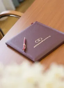 Eine lila Mappe mit einem goldenen Logo liegt auf einem Holztisch. Ein roter Stift liegt darauf. Auf der rechten Seite des Tisches befindet sich eine weiße Blume.