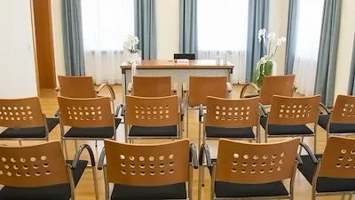 Ein leerer Konferenzraum mit braunen Stühlen, einem Holztisch und weißen Blumen in Vasen. Blaue Vorhänge hängen über den Fenstern.