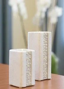Zwei weiße zylindrische Kerzenhalter stehen auf einem Holztisch, einer etwas kleiner als der andere. Beide haben eine strukturierte Oberfläche. Dahinter sind verschwommene weiße Blumen zu sehen.