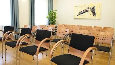 Ein leerer Konferenzraum mit Holzstühlen und einem schwarzen Tisch. Ein Gemälde ist an der Wand über den Stühlen angebracht. Ein Topf steht rechts.