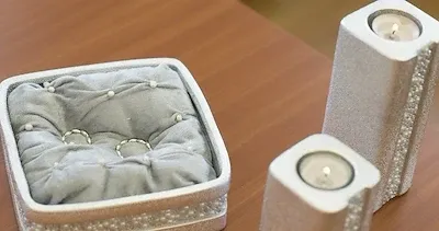 Eine kleine Box mit einem grauen Kissen enthält zwei silberne Ringe, und zwei zylindrische Kerzenhalter sind in der Nähe, einer angezündet und einer nicht.