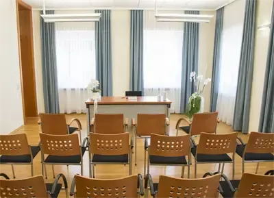 Ein leerer Konferenzraum mit einem Schreibtisch, Stühlen und einer weißen Wand. Ein Fenster mit blauen Vorhängen befindet sich hinter dem Schreibtisch.