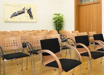 Ein leerer Besprechungsraum mit Holzböden und in Reihen angeordneten Stühlen. Ein modernes Gemälde schmückt die Wand über den Stühlen. Ein Topf mit einer Pflanze steht rechts neben einer Holztür.