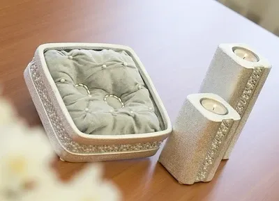 Eine dekorative Schachtel mit einem Kissen und zwei Kerzen auf einer Holzoberfläche. Die Schachtel ist mit Perlen verziert und enthält einen Ring.
