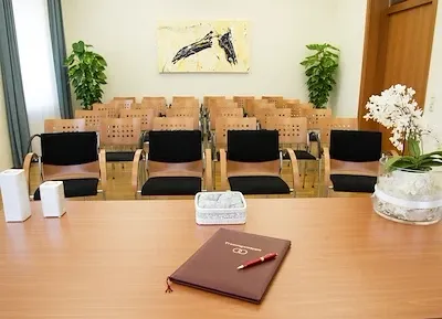 Ein leerer Konferenzraum mit Holzstühlen und -tischen. Auf dem Tisch stehen eine Blumenvase und ein Notizbuch, sowie einige Pflanzen im Hintergrund.