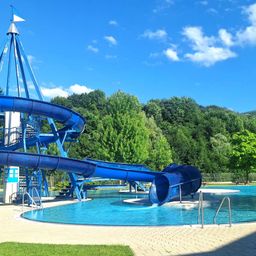 Eine große blaue Wasserrutsche mit einer Spiralform oben befindet sich in einem Außenpoolbereich, umgeben von üppigem grünen Bäumen und einem klaren blauen Himmel.