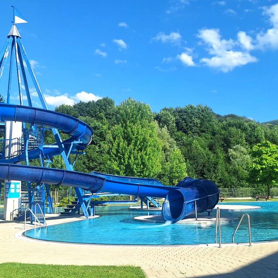 Eine große blaue Wasserrutsche mit einer Spiralform oben befindet sich in einem Außenpoolbereich, umgeben von üppigem grünen Bäumen und einem klaren blauen Himmel.