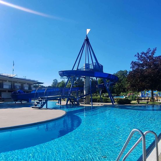 Eine blaue Wasserrutsche mit Spiraldesign steht neben einem großen Schwimmbecken an einem sonnigen Tag.