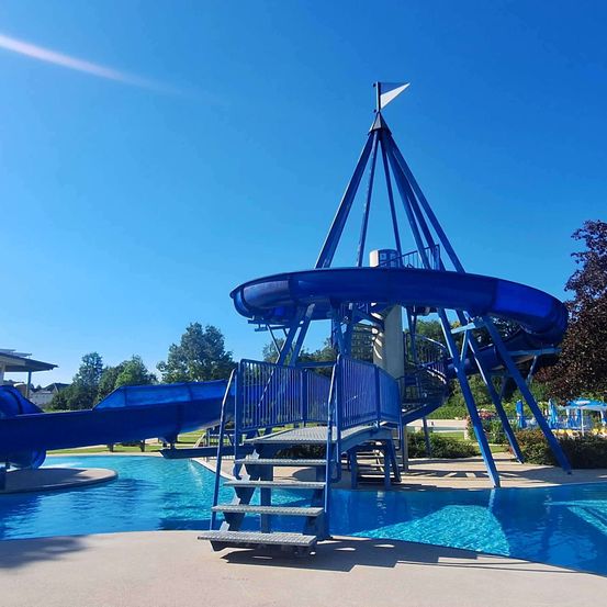 Eine blaue Wasserrutsche mit einer Wendeltreppe und einer Flagge auf der Spitze, neben einem Schwimmbecken unter einem klaren blauen Himmel.
