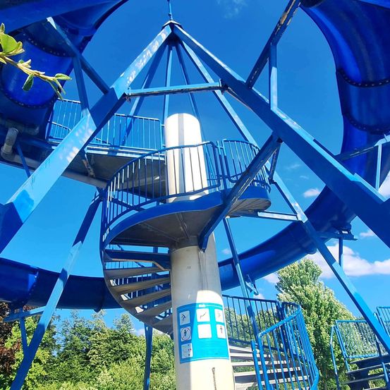 Eine blaue Spiralrutsche mit einer zentralen weißen Stützstruktur, umgeben von blauen Geländern und Stufen, unter einem klaren blauen Himmel.