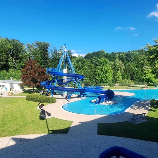 Eine große blaue Wasserrutsche spiralt in einen Swimmingpool, umgeben von üppigem Grün und einem klaren blauen Himmel.