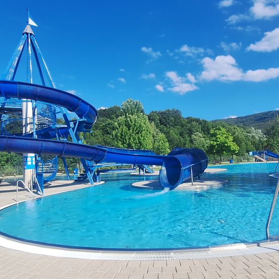 Eine große blaue Wasserrutsche spiralt in einen großen Swimmingpool, mit einem klaren blauen Himmel und grünen Bäumen im Hintergrund.