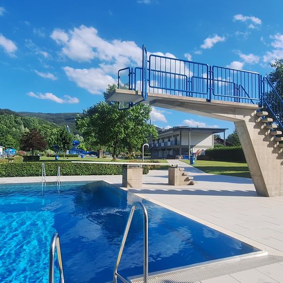 Ein Freibad mit Sprungbrett, blauen Geländern und einem klaren blauen Himmel darüber.
