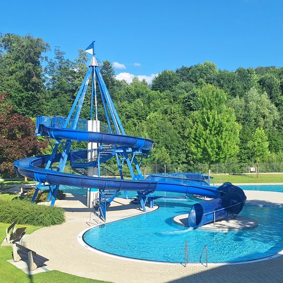 Eine blaue Wasserrutsche mit einer Spiralform befindet sich in einem Außenpoolbereich, umgeben von üppigem Grün und einem klaren blauen Himmel.