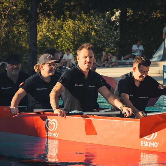 Vier Männer in schwarzen Shirts rudern mit einem orangefarbenen Eskimo-Boot im Pool, mit Zuschauern im Hintergrund.